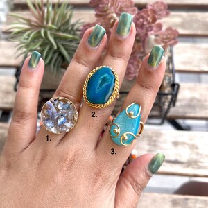 May include: Three gemstone rings are shown on a hand. Ring 1 is a round, gold-toned ring with a light blue and white stone. Ring 2 is an oval, gold-toned ring with a blue stone. Ring 3 is a teardrop-shaped, gold-toned ring with a blue stone.