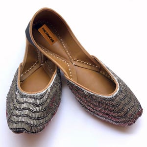 Peut inclure: Une paire de ballerines argent&eacute;es &agrave; perles avec un motif ondul&eacute;. Les chaussures ont une semelle int&eacute;rieure en cuir marron et un talon noir. La marque "THE ENACHE" est imprim&eacute;e sur une petite &eacute;tiquette marron &agrave; l'int&eacute;rieur de la chaussure.