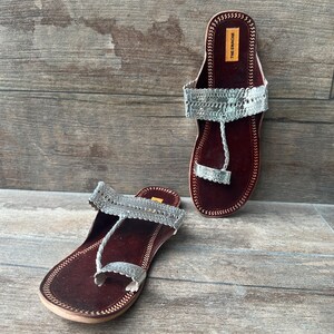 May include: A pair of silver and brown leather sandals with a toe ring. The sandals have a decorative silver design on the straps and are labeled "The Enachie".