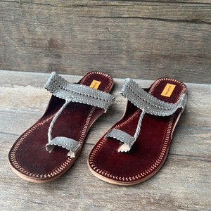 May include: A pair of silver leather sandals with intricate cutouts and a braided strap. The sandals have a maroon leather sole and are made by "The Ethnic" brand.