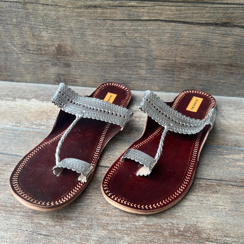 May include: A pair of silver leather sandals with intricate cutouts and a braided strap. The sandals have a maroon leather sole and are made by "The Ethnic" brand.