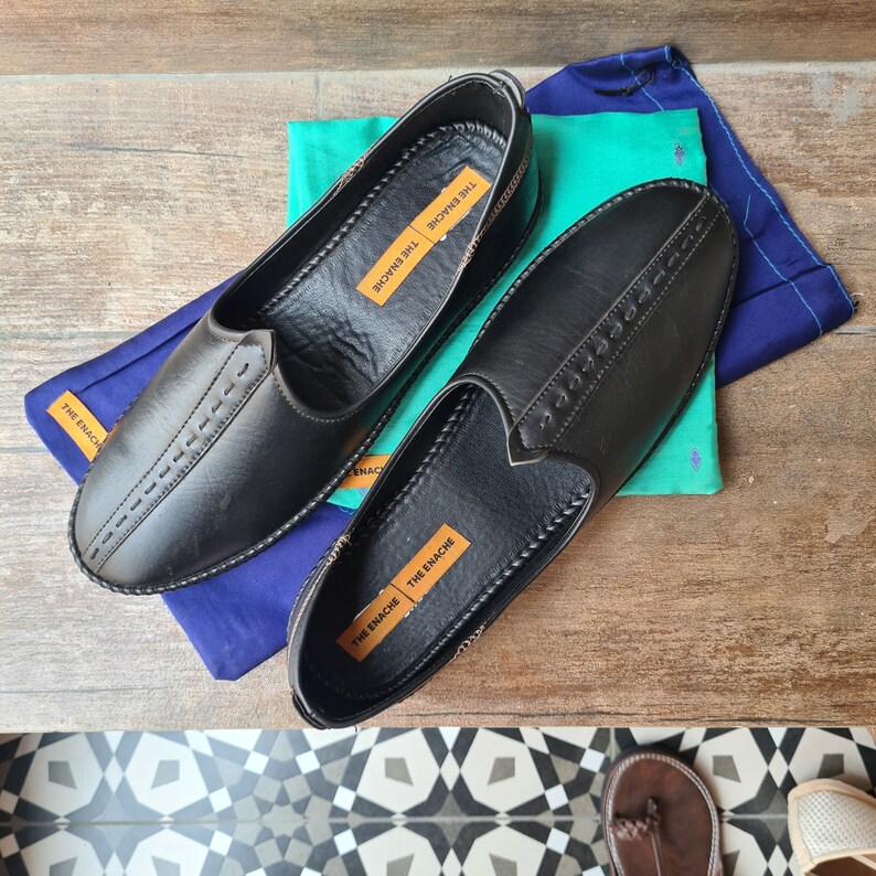 Op de afbeelding: Een paar zwarte leren instappers met decoratieve stiksels. De schoenen liggen op een houten oppervlak met blauwe en groene stof. Het label "THE ENACHE" is zichtbaar. De schoenen zijn een klassiek model.