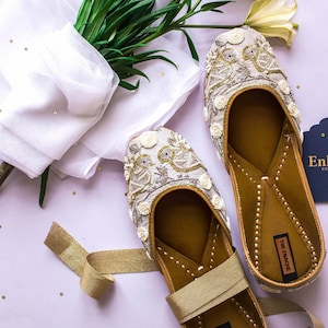 May include: Beige embroidered flats with bird and floral patterns. The flats have a gold strap and a brown sole. A white scarf and greenery are in the background. The flats are labelled "The Enache".