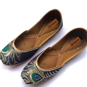 Könnte beinhalten: Ein Paar marineblaue und lila Ballerinas mit Pfauenfedern-Stickerei. Die Schuhe haben eine braune Lederinnenseite und einen goldenen Besatz. Der Text "THE ENACHE" ist auf einem braunen Etikett im Inneren jedes Schuhs aufgedruckt.