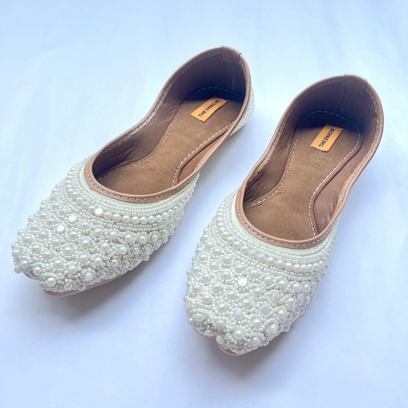 May include: A pair of white flat shoes with pearl embellishments. The shoes have a brown leather insole with the text "The Encore" printed on them.