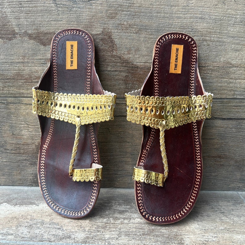May include: A pair of brown leather sandals with gold embellishments. The sandals have a toe loop and an ankle strap. The brand name "THE ENACHE" is printed on the straps.
