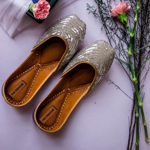 May include: A pair of silver sequined slip-on shoes with tan leather interiors and soles. The shoes have a pointed toe and are decorated with the text "THE ENACHE" on the inner sole. The shoes are displayed on a light purple surface with floral accents.