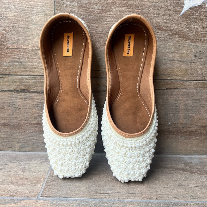 May include: A pair of white flat shoes with pearl embellishments. The shoes have a brown leather insole with the text "THE ENACHE" printed on them.