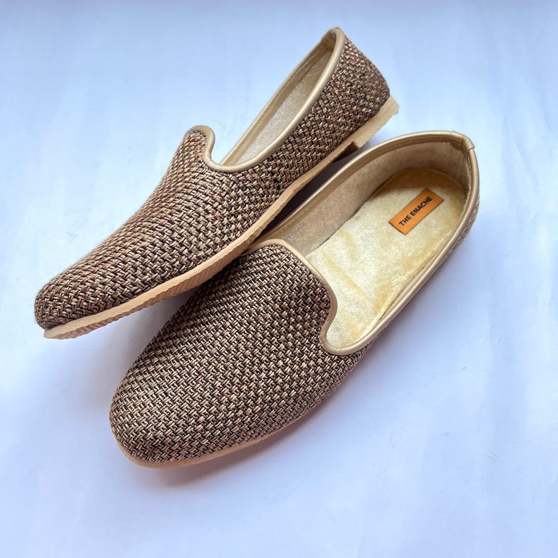 Op de afbeelding: Een paar bruine geweven slippers met een gouden glans. De slippers hebben een zacht, gevoerd voetbed en een comfortabele pasvorm. De merknaam "THE EMACHE" is zichtbaar op de binnenzool van een slipper.