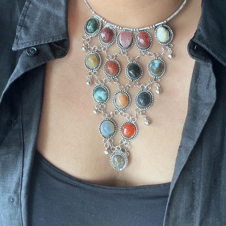 May include: A silver necklace with a cascade of colorful gemstones. The necklace features a variety of oval-shaped stones in shades of green, red, pink, yellow, and blue. The stones are set in a silver filigree design.