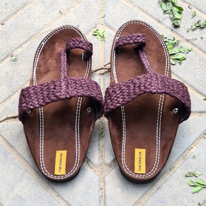 May include: A pair of brown leather sandals with a purple braided strap. The sandals have a toe loop and the brand name "THE ENACHE" is printed on the sole.