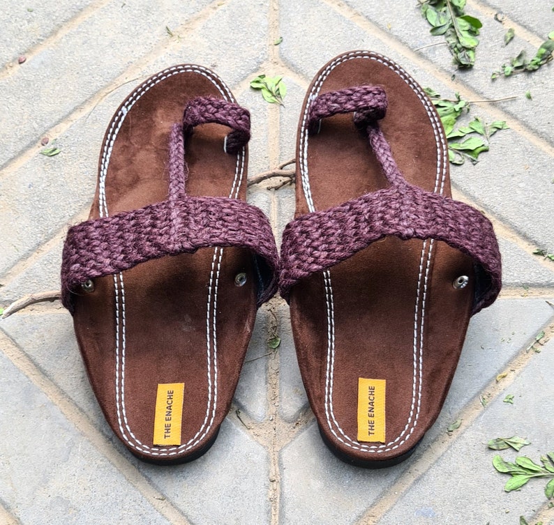 May include: A pair of brown leather sandals with a purple braided strap. The sandals have a toe loop and the brand name "THE ENACHE" is printed on the sole.