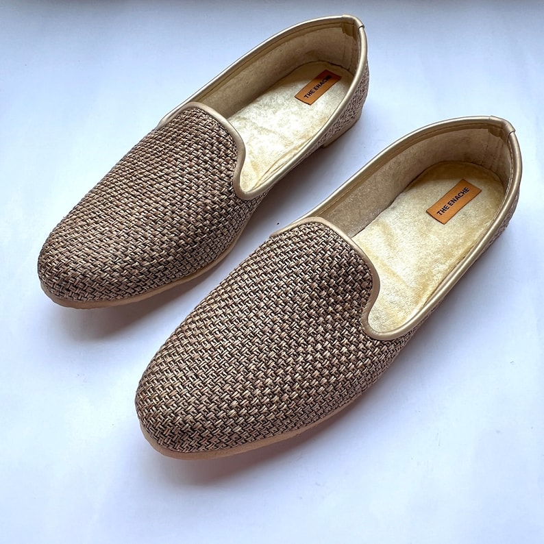 Op de afbeelding: Een paar gouden geweven slippers met een zachte, pluche voering. De slippers hebben de tekst "THE  ENACHE" gedrukt aan de binnenkant.