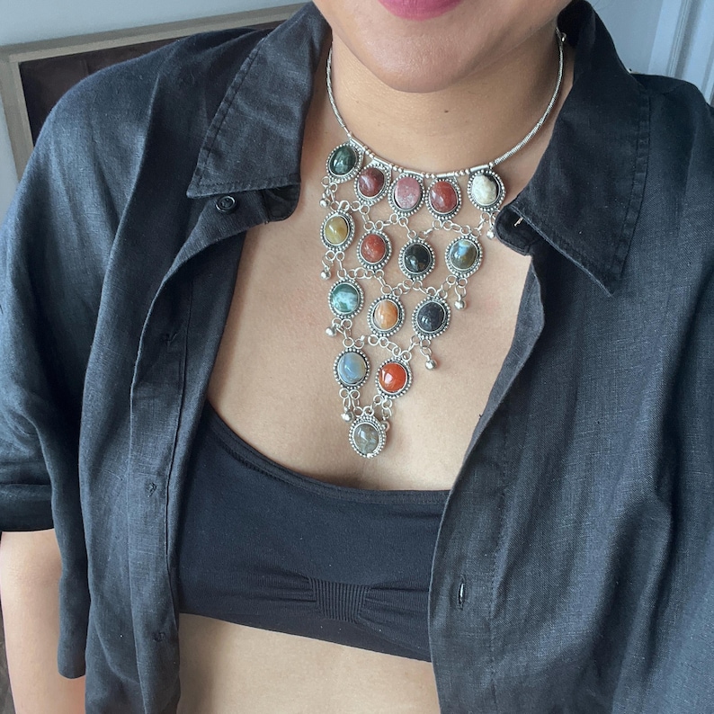 May include: A silver-toned necklace with multiple colorful gemstone pendants. The necklace features a variety of round stones in shades of red, green, pink, and black. The necklace is worn over a black top and a dark gray button-down shirt.