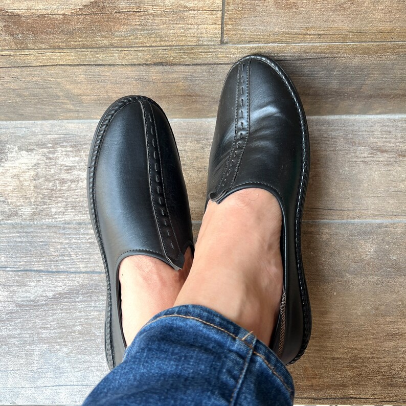 Op de afbeelding: Een paar zwarte leren instappers. De schoenen hebben een ronde neus en een gestikt detail aan de bovenkant. De zolen zijn zwart met een textuurpatroon. De schoenen worden van bovenaf getoond.