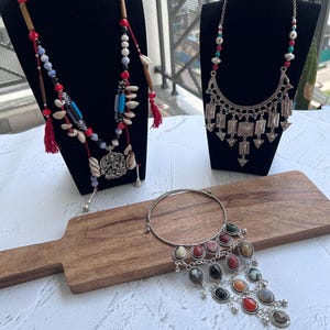 May include: Three necklaces on black stands and a wooden board. One necklace has red tassels, wooden beads, and shell accents. Another features a silver pendant with geometric shapes. The third has a silver band with colorful stone pendants.