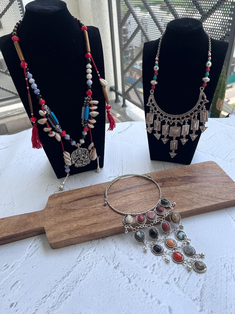 May include: Three necklaces on black stands and a wooden board. One necklace has red tassels, wooden beads, and shell accents. Another features a silver pendant with geometric shapes. The third has a silver band with colorful stone pendants.