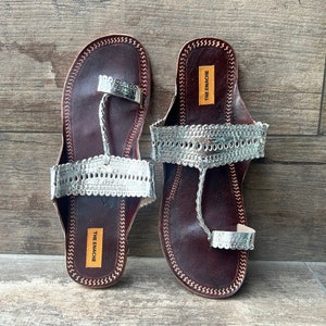 May include: A pair of brown leather sandals with silver embellishments. The sandals have a toe loop and an ankle strap. The brand name "THE ENACHE" is printed on the sole of each sandal.
