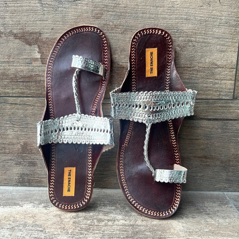 May include: A pair of brown leather sandals with silver embellishments. The sandals have a toe loop and an ankle strap. The brand name "THE ENACHE" is printed on the sole of each sandal.