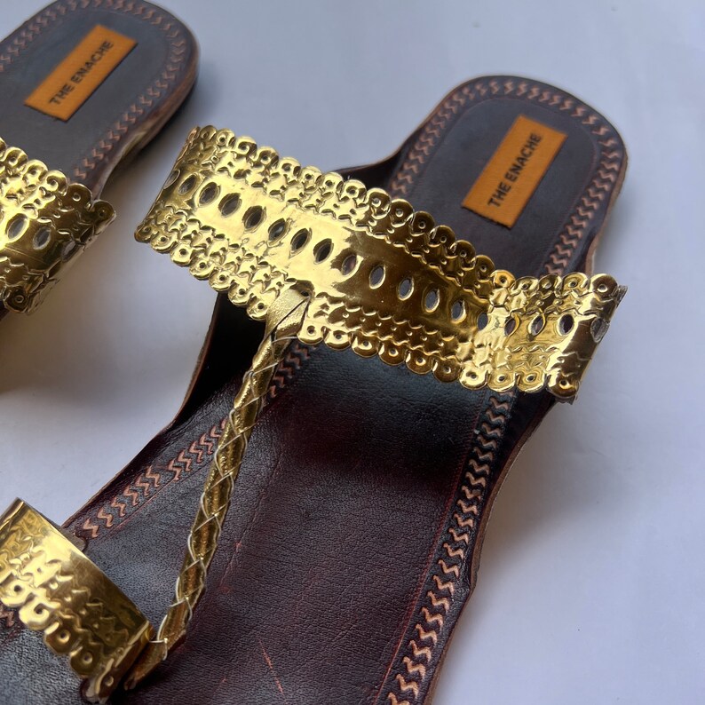 May include: Brown leather sandals with a gold metal strap and the text "THE ENACHE" on the strap.