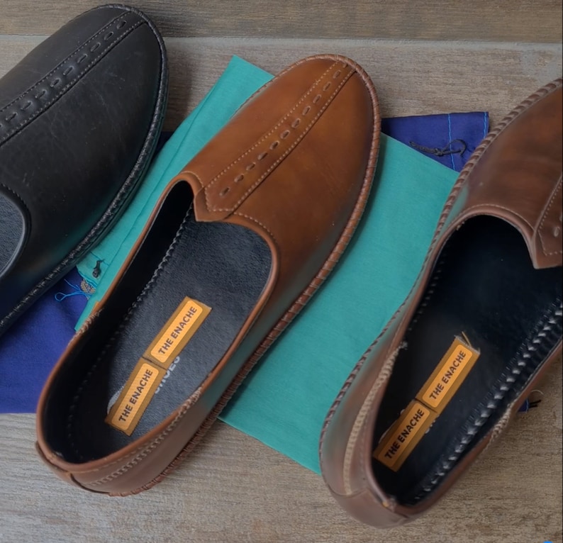 Op de afbeelding: Een close-up van drie leren loafers in zwart en bruin. De bruine loafers staan centraal, met een gestikt ontwerp aan de bovenkant. Elke schoen heeft een zwart interieur en een label met de tekst "THE ENACHE". De schoenen liggen op een houten oppervlak.
