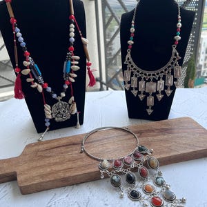 May include: Three necklaces on display. One features red tassels, wooden beads, and shells. Another has a silver pendant with geometric shapes. The third is a silver choker with colorful oval stones and dangling charms. All are displayed on black mannequins and a wooden board.