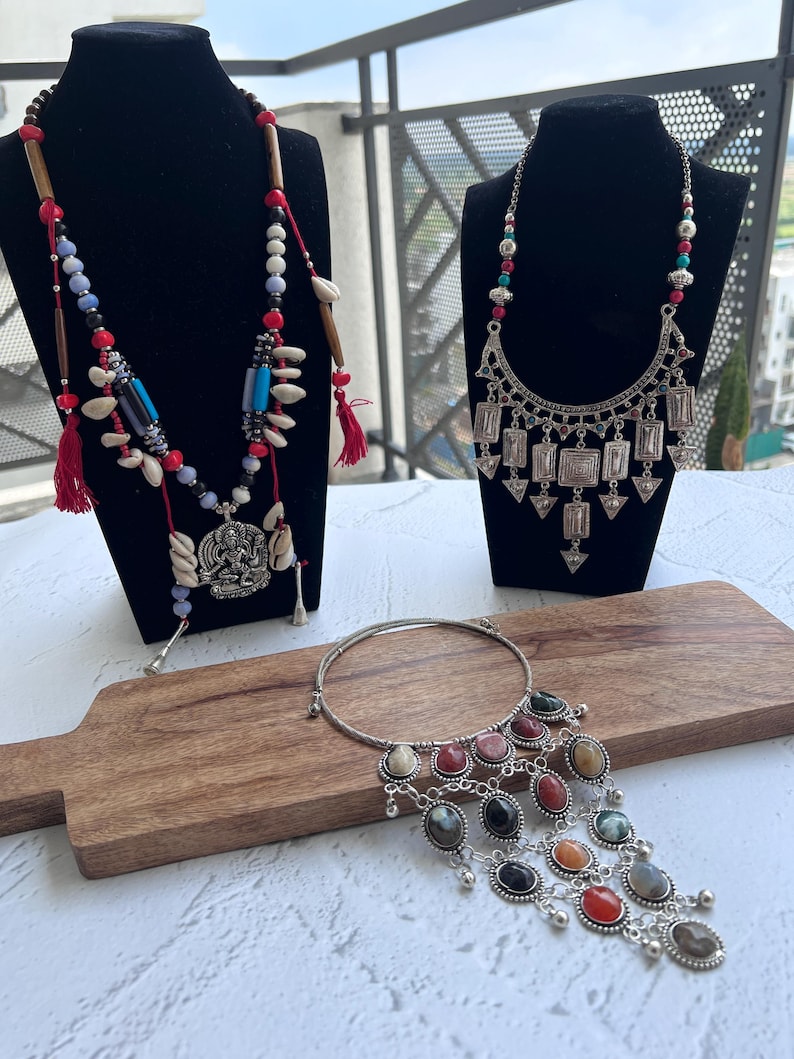 May include: Three necklaces on display. One features red tassels, wooden beads, and shells. Another has a silver pendant with geometric shapes. The third is a silver choker with colorful oval stones and dangling charms. All are displayed on black mannequins and a wooden board.