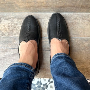 Op de afbeelding: Een paar zwarte leren instappers met een ronde neus en decoratieve stiksels. De schoenen worden gedragen met een blauwe spijkerbroek. De foto is van bovenaf genomen.