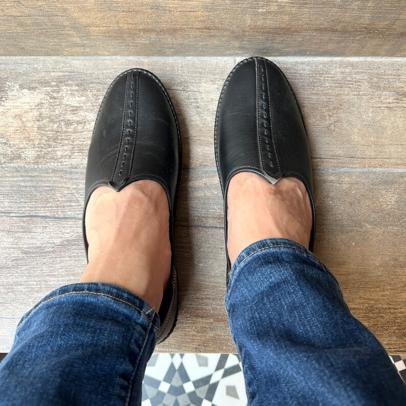 Op de afbeelding: Een paar zwarte leren instappers met een ronde neus en decoratieve stiksels. De schoenen worden gedragen met een blauwe spijkerbroek. De foto is van bovenaf genomen.