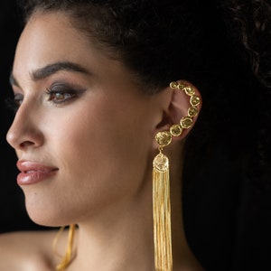 May include: A gold ear cuff with a row of round, faceted stones. A long, gold chain dangles from the bottom of the cuff, forming a tassel.