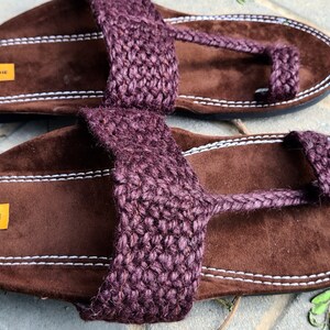 May include: A pair of brown leather sandals with a purple jute strap and a toe loop. The sandals have a white stitching detail and a label that reads "THE ENACHE".