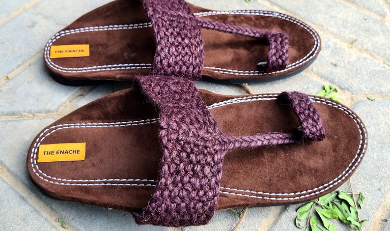 May include: A pair of brown leather sandals with a purple jute strap and a toe loop. The sandals have a white stitching detail and a label that reads "THE ENACHE".