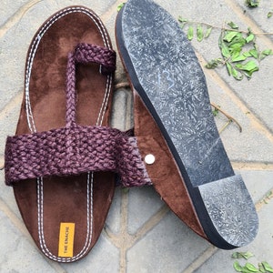 May include: Brown leather sandals with a woven strap and a rubber sole. The sandals have a toe loop and the brand name "THE ENACHE" is printed on the sole.