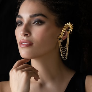 Puede incluir: Una mujer lleva un pendiente de oreja dorado con piedras negras y una cadena colgante. El pendiente de oreja tiene forma de flor.