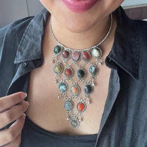 May include: Silver-toned necklace featuring a cascade of colorful, round gemstones. The necklace has a delicate chain and intricate metalwork, with a variety of stone colors including green, red, pink, and orange. Small dangling accents add movement to the design.