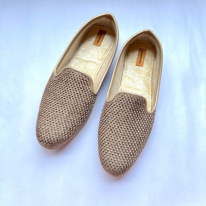 Op de afbeelding: Een paar gouden geweven slippers met de tekst "The Emach&eacute;" aan de binnenkant van elke schoen.