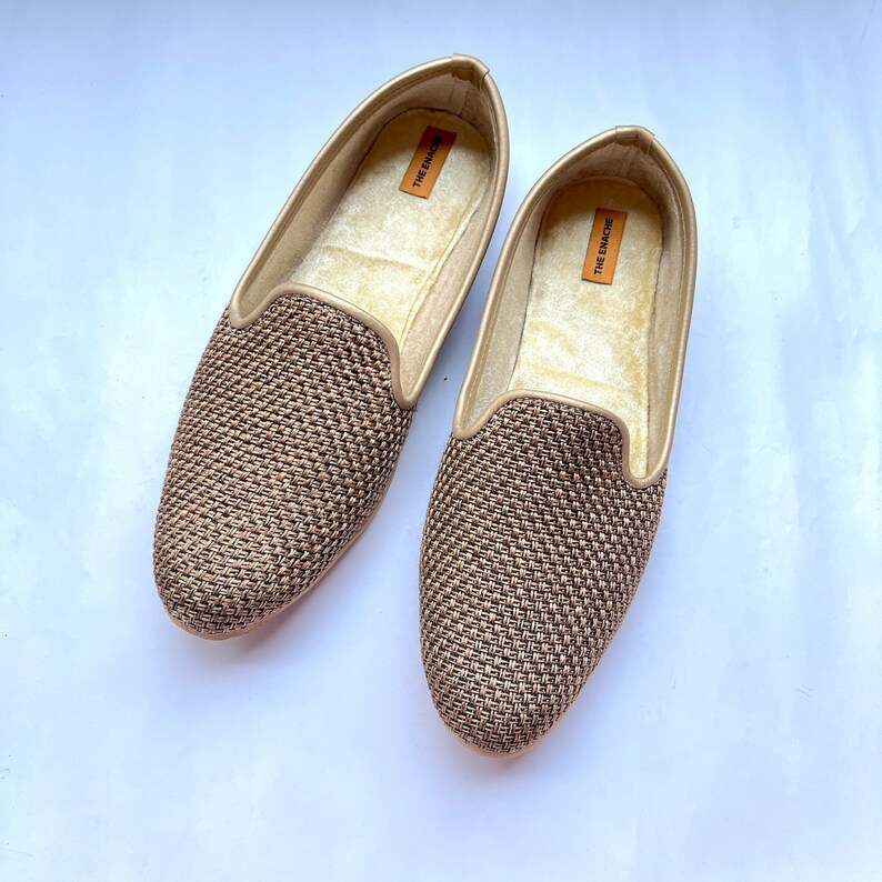 Op de afbeelding: Een paar gouden geweven slippers met de tekst "The Emach&eacute;" aan de binnenkant van elke schoen.