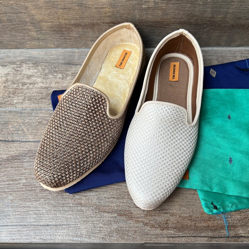 Op de afbeelding: Twee paar geweven leren slippers, een bruine en een witte, met de merknaam "THE ENACRE" gedrukt aan de binnenkant van elke slipper.