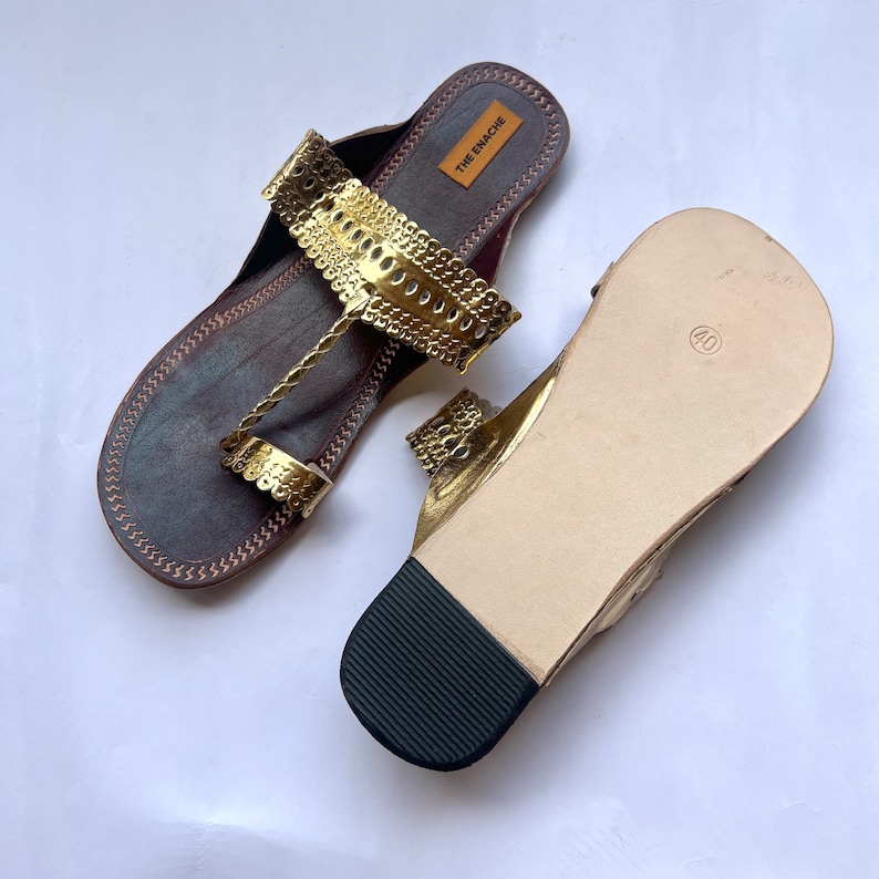 May include: A pair of gold leather sandals with a decorative cut-out design. The sandals have a brown leather sole and a black rubber heel. The sandals are labeled "THE GMACHE" on the insole.