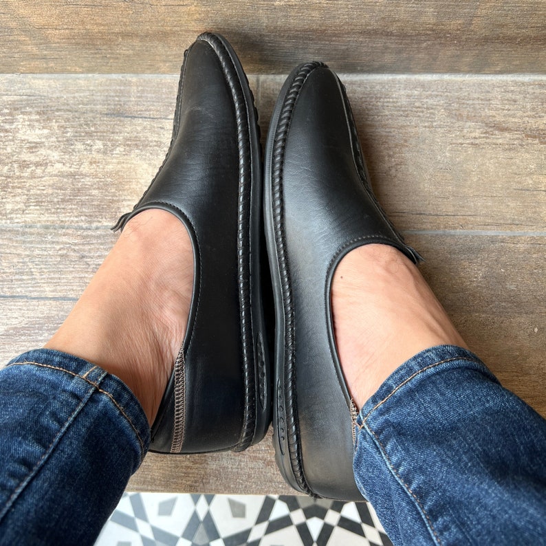 Op de afbeelding: Een paar zwarte leren instappers met een ronde neus en gestikte details. De schoenen hebben een laag profiel en worden van bovenaf getoond, met de voeten van de drager zichtbaar. De schoenen worden gecombineerd met een blauwe spijkerbroek.
