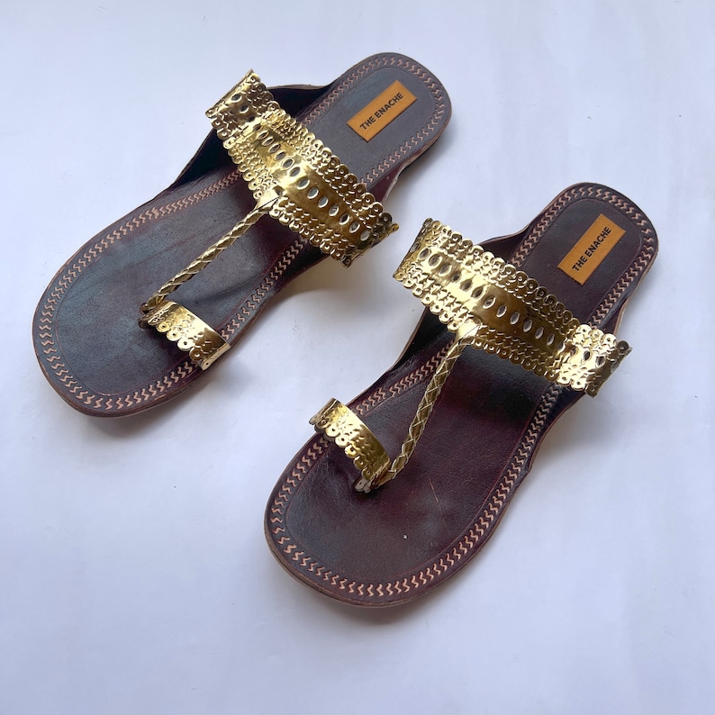 May include: A pair of brown leather sandals with gold metal embellishments. The sandals have a toe loop and an ankle strap. The brand name "THE ENACHE" is printed on the straps.