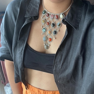 May include: A silver necklace with multiple colorful gemstone pendants. The necklace features a variety of round gemstones in shades of red, green, yellow, and blue. The necklace is worn over a black top and a dark gray button-down shirt.