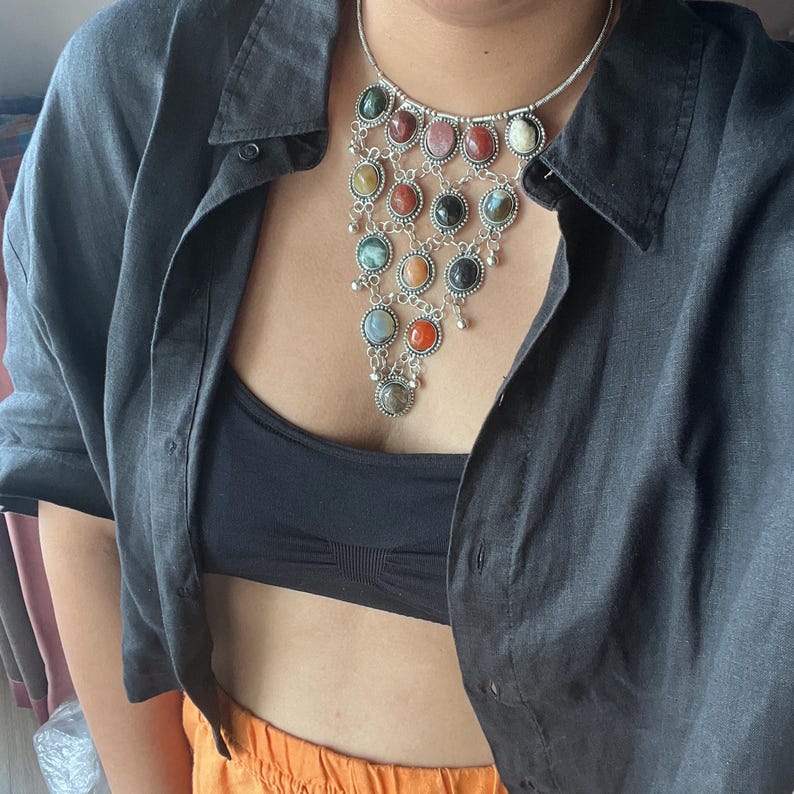 May include: A silver necklace with multiple colorful gemstone pendants. The necklace features a variety of round gemstones in shades of red, green, yellow, and blue. The necklace is worn over a black top and a dark gray button-down shirt.
