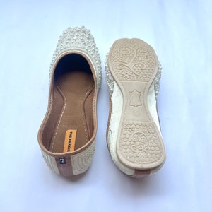 May include: A pair of white flat shoes with pearl embellishments. The shoes have a brown leather insole with the text "THE ENACHI" and the size "12" printed on them. The shoes are made in India.