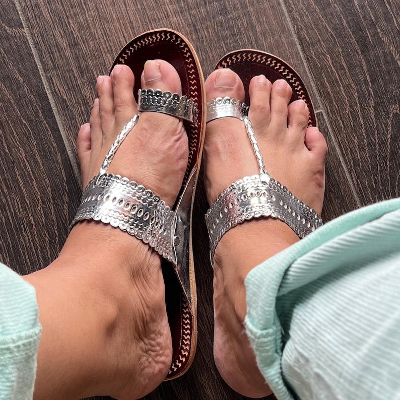 May include: A pair of silver and brown leather sandals with decorative silver embellishments. The sandals have a toe strap and an ankle strap.