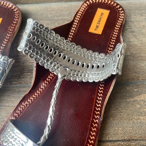 May include: Silver leather sandals with a decorative strap and a brown leather sole. The sandals have a brand label that says "THE EMACHE".