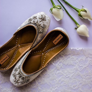 May include: A pair of white, embellished slip-on shoes with floral and crystal accents. The shoes have brown leather insoles and are labeled "THE ENACHE". White roses and lace are in the background.