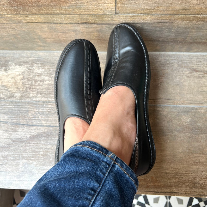 Op de afbeelding: Een paar zwarte leren instappers met decoratieve stiksels. De schoenen hebben een ronde neus en een laag profiel. De afbeelding toont de schoenen vanuit een vogelperspectief, met de voeten van een persoon die een blauwe spijkerbroek draagt.