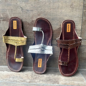May include: Three pairs of brown leather sandals with decorative gold, silver, and bronze embellishments. The sandals have a toe loop and an ankle strap. The brand name "THE ENACHE" is printed on the straps.