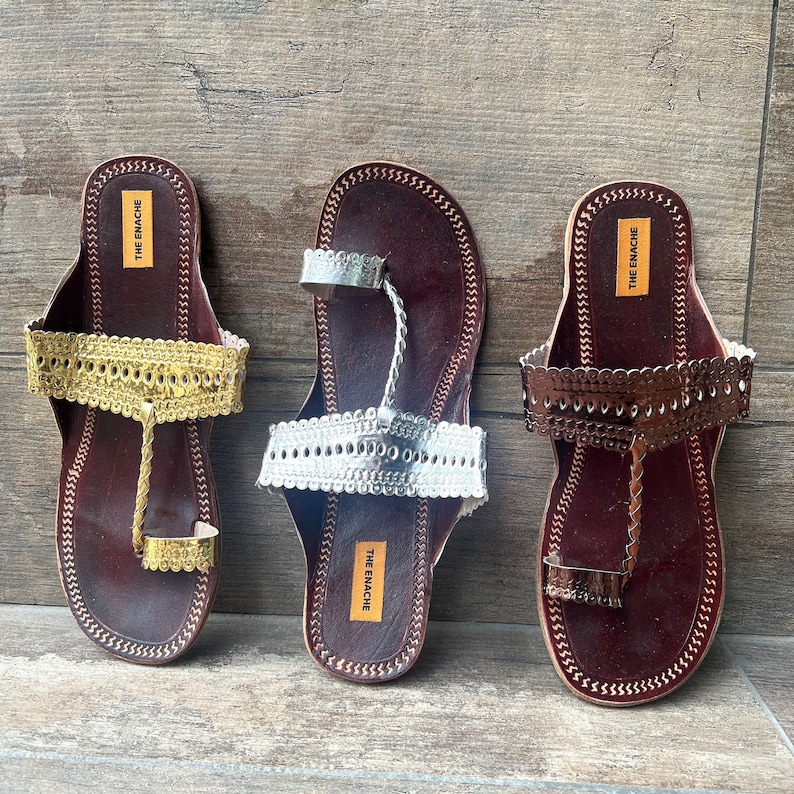 May include: Three pairs of brown leather sandals with decorative gold, silver, and bronze embellishments. The sandals have a toe loop and an ankle strap. The brand name "THE ENACHE" is printed on the straps.